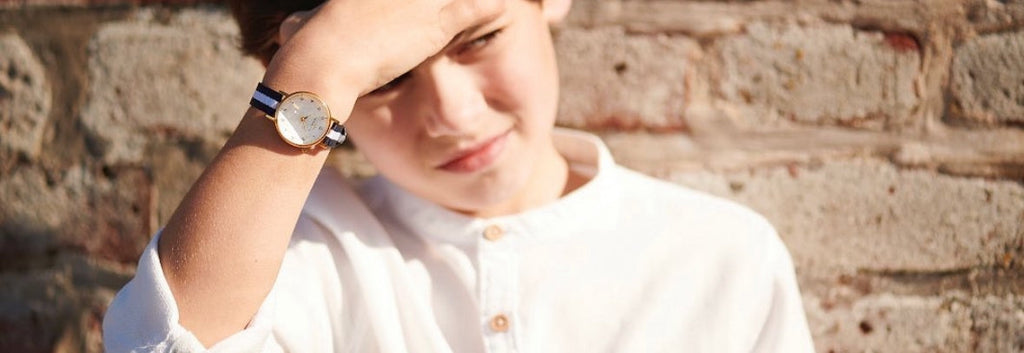 Pojke mot väggen i Torekov med sin analoga barnklocka i guld och mjuk nato i marint och vitt - Torekovs klockor